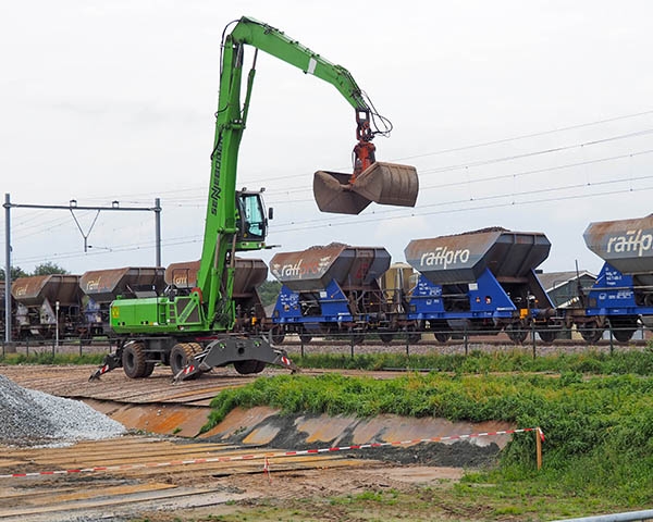 Groene overslagkraan met grijperbak boven een rij ballastwagons langs het spoor
