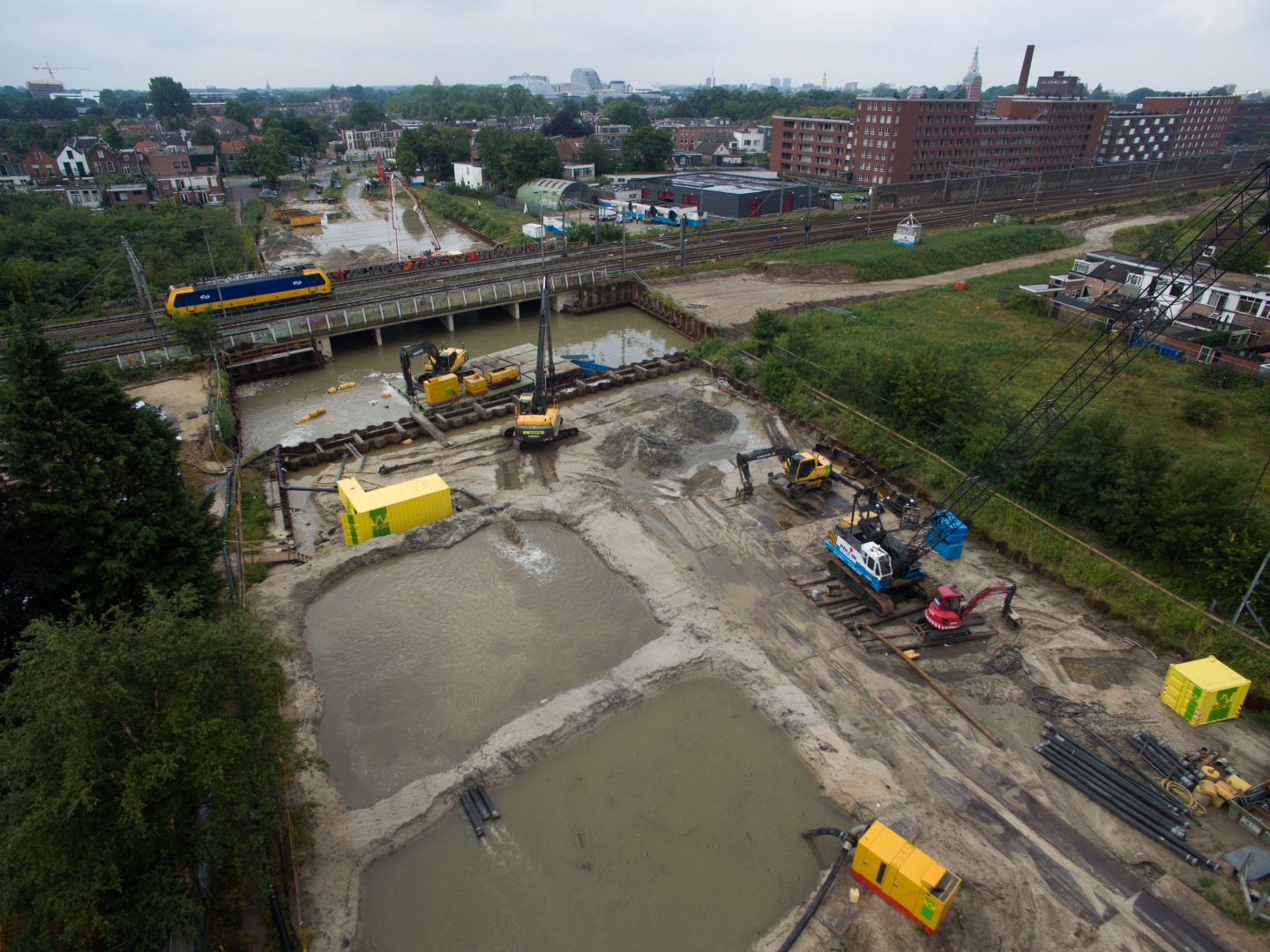 Luchtfoto van twee natte bouwkuipen naast een spoorlijn, met kranen op de damwand