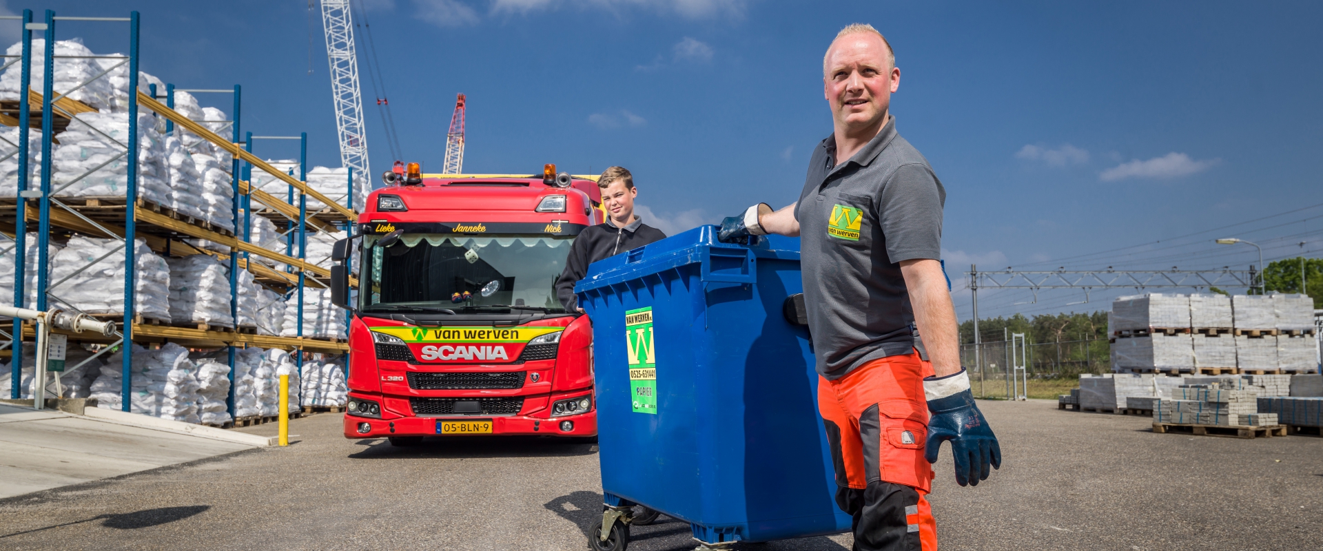Twee medewerkers van Van Werven rollen een blauwe afvalcontainer over het terrein, met daarachter een vrachtwagen van Van Werven