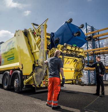 Een gele inzamelwagen van Van Werven leegt een blauwe rolcontainer; twee medewerkers kijken toe