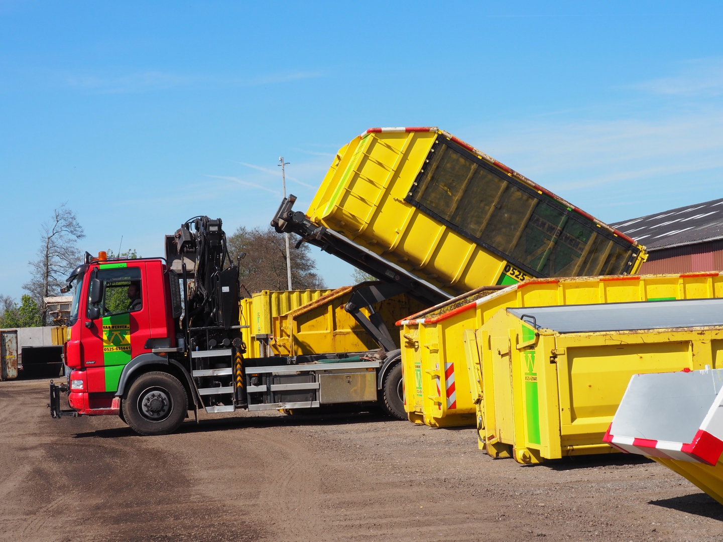 Vrachtwagen van Van Werven zet met een kraan een gele afzetcontainer neer op een terrein met meer containers