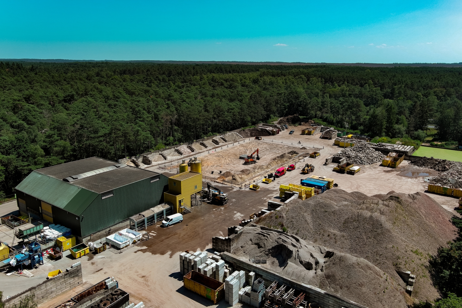 Luchtfoto van de recyclinglocatie tussen de bossen, met grondbergen, gele containers en de groene loods