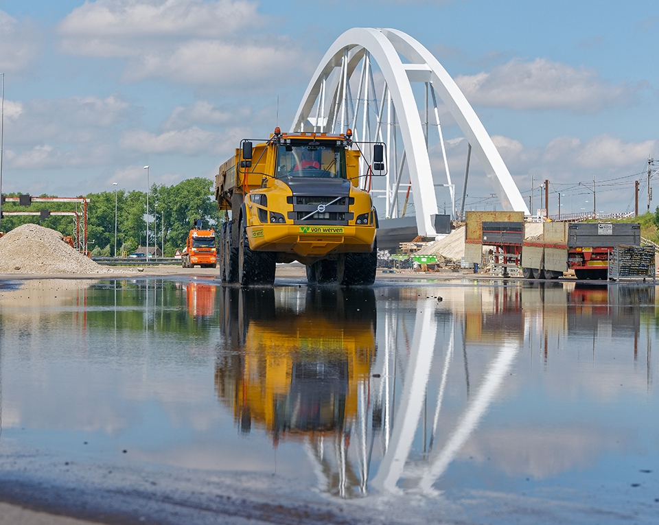 Knikdumper van Van Werven rijdt door een plas water voor de witte boogbrug van een nieuw viaduct