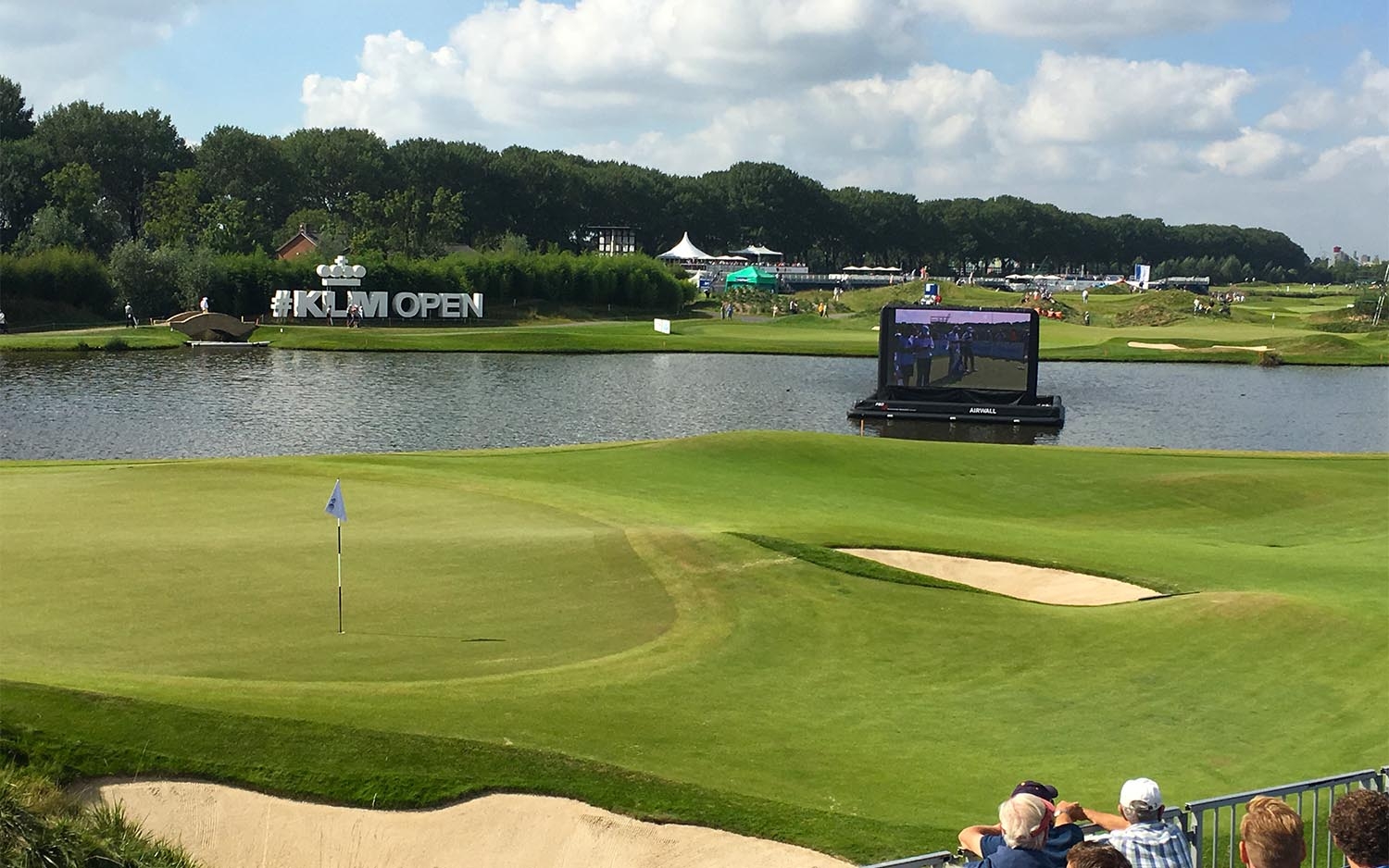 Green van een golfbaan tijdens het KLM Open, met vlag en bunker in de voorgrond en een drijvend scherm op het water erachter
